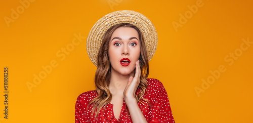 Surprised, shocked young woman in a straw hat, in a red summer dress, red lipstick looks directly at the camera. Portrait of a shocked girl in summer clothes on an orange background, sale concept