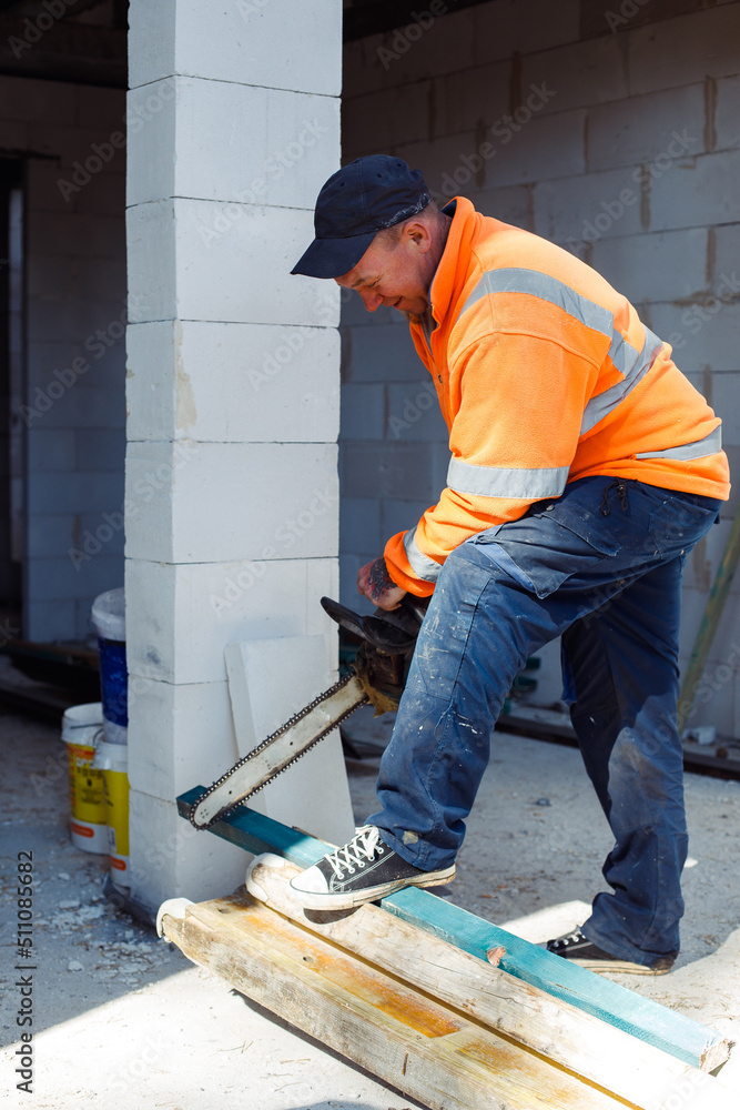 Vertical smiling worker man in orange protective coverall using chainsaw cutting wood piece and metal tubes. Build house Vertical smiling worker man in orange protective coverall using chainsaw cutting wood piece and metal tubes. Build house