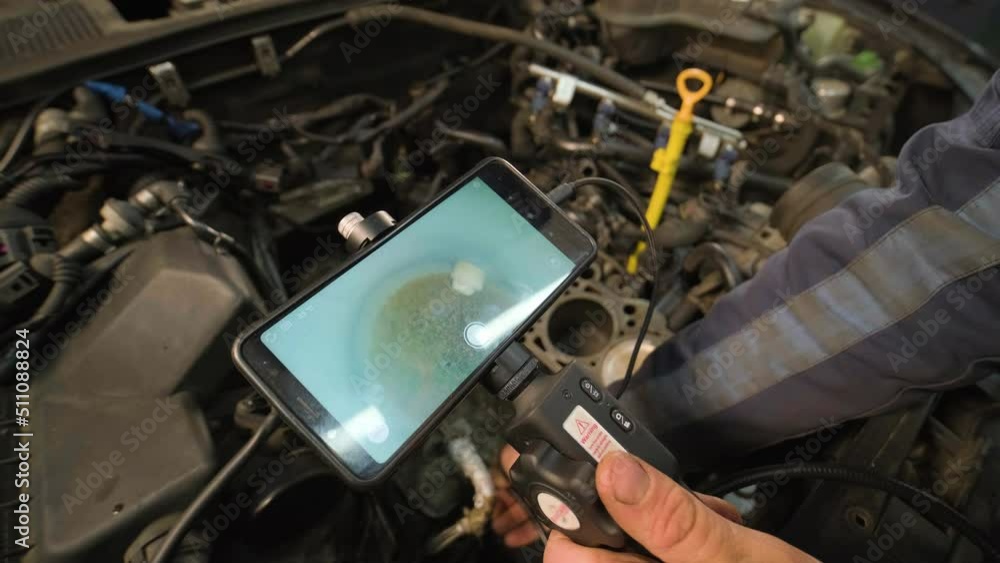Hands of car mechanic check the vehicle engine with technical endoscope with rotary camera and mobile phone. Repair of engine head and valves.