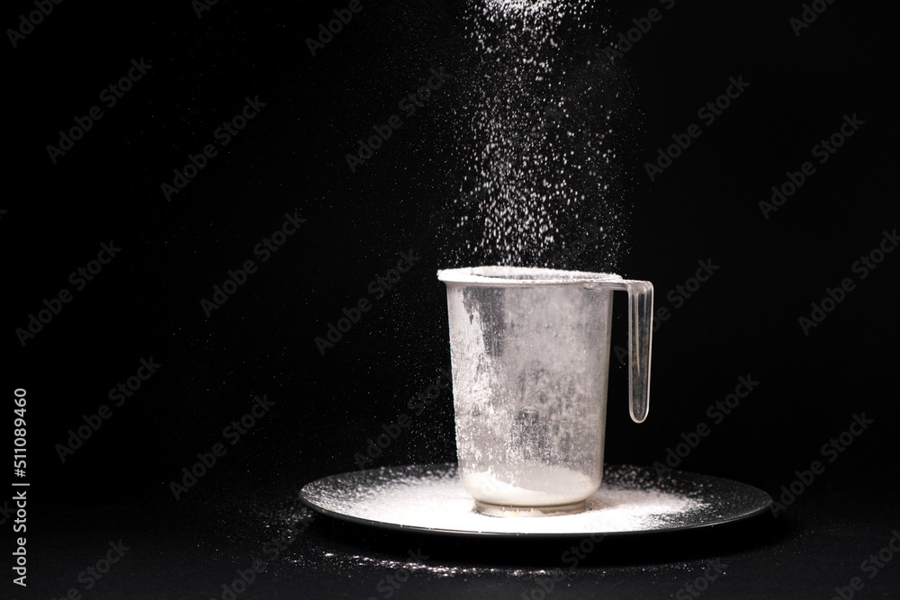 White wheat flour is poured into a measuring cup on a black background. Cooking and baking, a place for text. White wheat flour is poured into a measuring cup on a black background. Cooking and baking, a place for text.