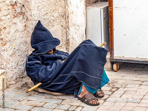 Elderly gentleman rests against an ancient wall of the medina in his djellaba, a royal blue traditional robe with pointed hood.