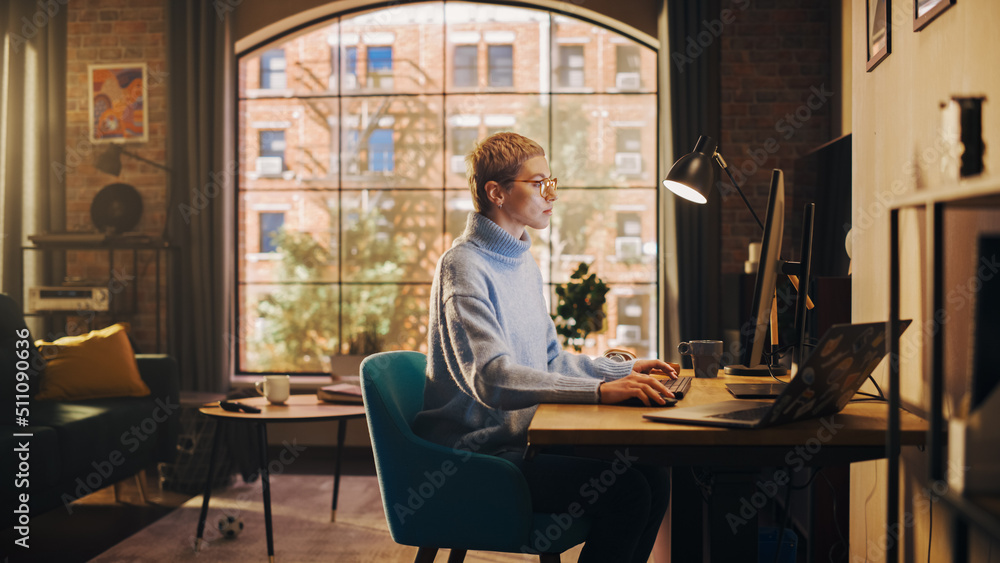 Young Attractive Woman Working from Home on Desktop Computer in Sunny Stylish Loft Apartment. Creative Designer Wearing Cozy Blue Sweater and Glasses. Urban City View from Big Window. Young Attractive Woman Working from Home on Desktop Computer in Sunny Stylish Loft Apartment. Creative Designer Wearing Cozy Blue Sweater and Glasses. Urban City View from Big Window.