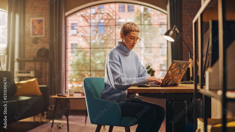 Young Diverse Woman Working from Home on Laptop Computer with Stickers in Sunny Stylish Loft Apartment. Creative Designer Wearing Cozy Blue Sweater and Glasses. Urban City View from Big Window.