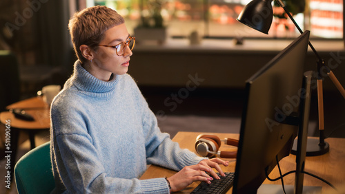 Young Beautiful Woman Sitting Down to Work on Desktop Computer in Sunny Stylish Loft Apartment. Creative Designer Wearing Cozy Blue Sweater and Glasses. Urban City View from Big Window.