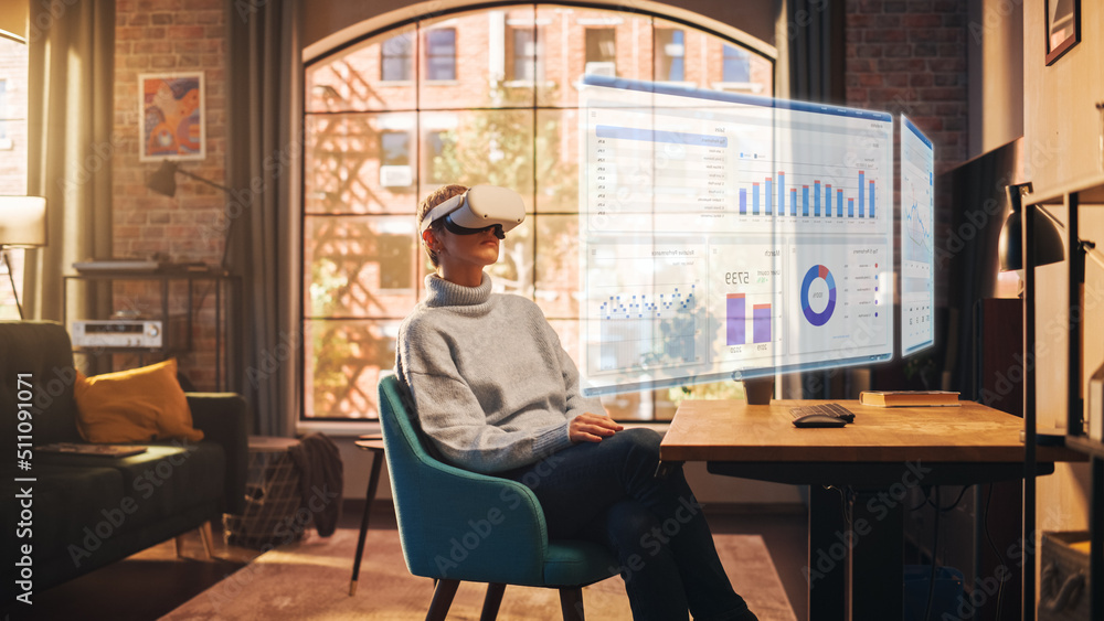 Young Female Employee with Short Hair Works From Home in Virtual Reality Goggles. She is Wearing a Modern Headset and Using Controllers to Scroll Through Graphs. Stylish Apartment with Big Window. Young Female Employee with Short Hair Works From Home in Virtual Reality Goggles. She is Wearing a Modern Headset and Using Controllers to Scroll Through Graphs. Stylish Apartment with Big Window.