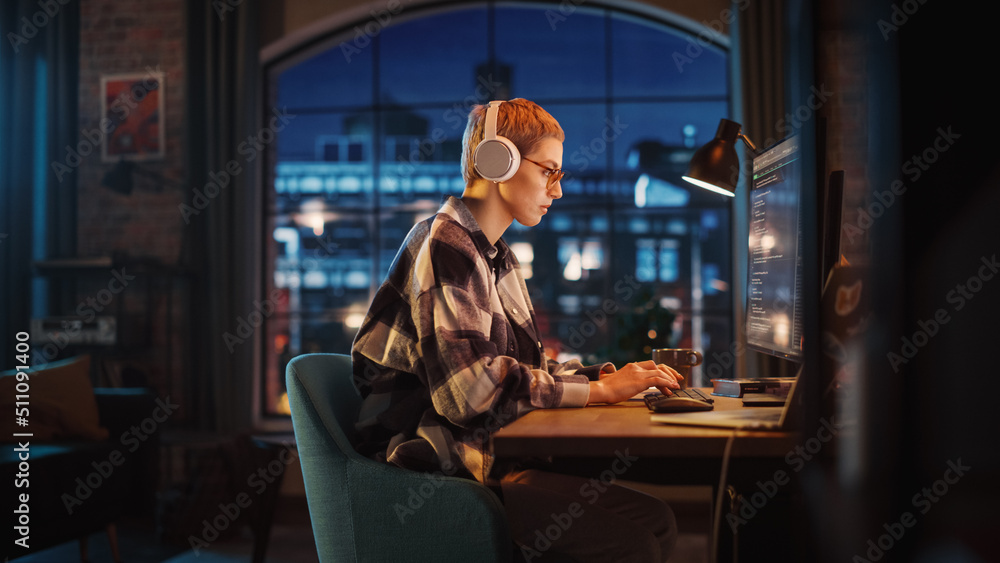 Young Woman Writing Code on Desktop Computer in Stylish Loft Apartment in the Evening. Creative Female Wearing Headphones, Working from Home on Software Development. Urban City View from Big Window.