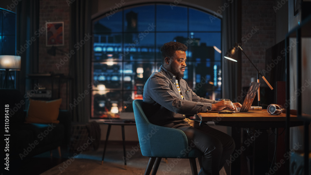 Young Handsome African American Man Working from Home on Laptop Computer in Stylish Loft Apartment at Night. Creative Male Checking Social Media, Browsing Internet. Urban City View from Big Window.