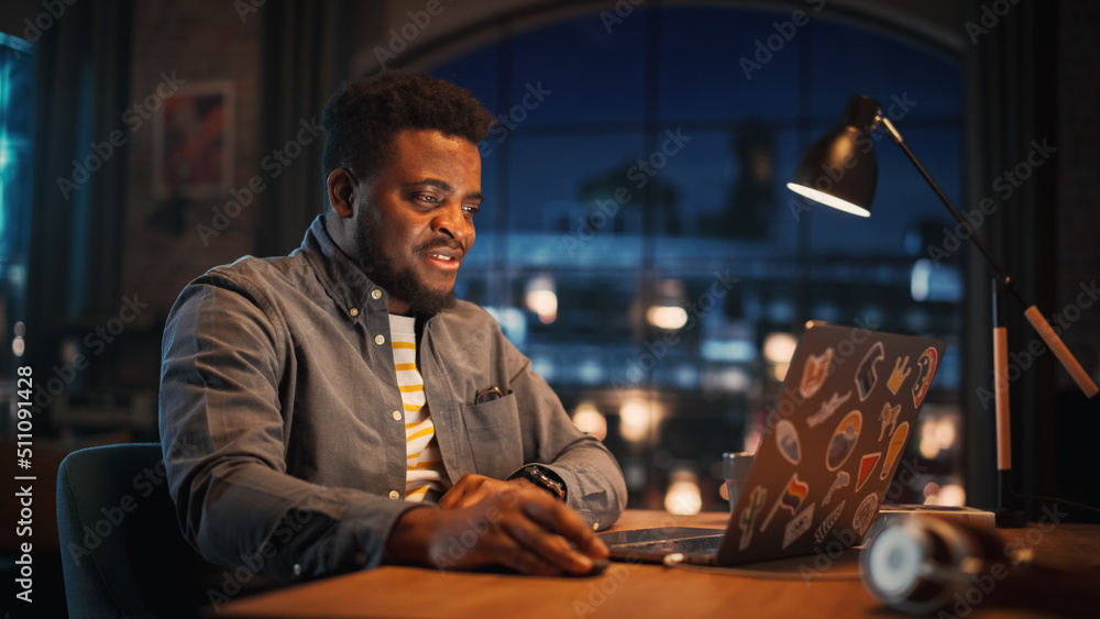 Young Handsome Black Man Working from Home on Laptop Computer in Stylish Loft Apartment in the Evening. Creative Male Gets a Great Idea while Checking Social Media. Urban City View from Big Window. Young Handsome Black Man Working from Home on Laptop Computer in Stylish Loft Apartment in the Evening. Creative Male Gets a Great Idea while Checking Social Media. Urban City View from Big Window.