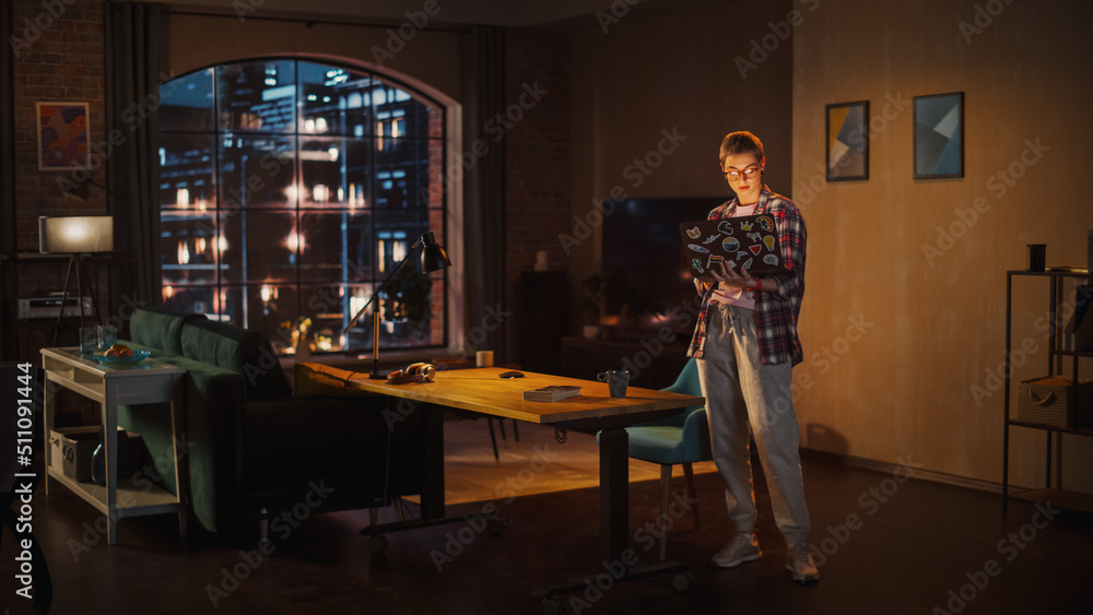 Young Beuatiful Women Working from Home, Standing And Holding Laptop Computer in Stylish Loft Apartment in the Evening. Creative Female Wearing Cozy Casual Outfit. Urban City View from Big Window.