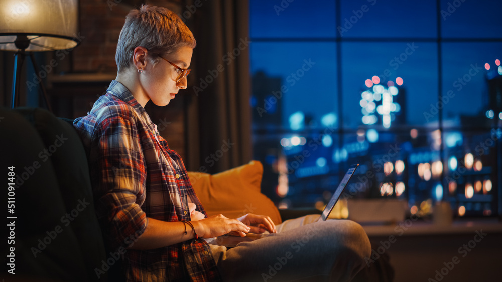 Young Beautiful Woman Sitting on Couch, Using Laptop Computer in Stylish Loft Apartment in the Evening. Creative Female Smiling, Checking Social Media, Stretching. Urban City View from Big Window.