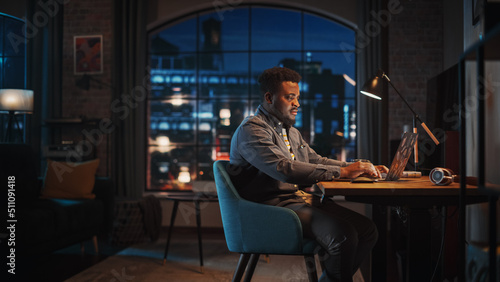 Young Handsome African American Man Working from Home on Laptop Computer in Stylish Loft Apartment at Night. Creative Male Checking Social Media, Browsing Internet. Urban City View from Big Window.