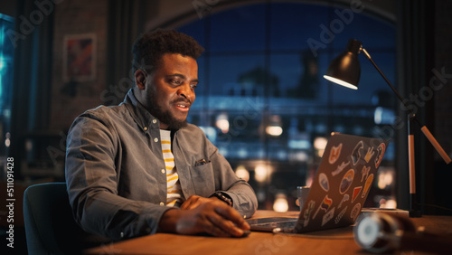 Young Handsome Black Man Working from Home on Laptop Computer in Stylish Loft Apartment in the Evening. Creative Male Gets a Great Idea while Checking Social Media. Urban City View from Big Window.