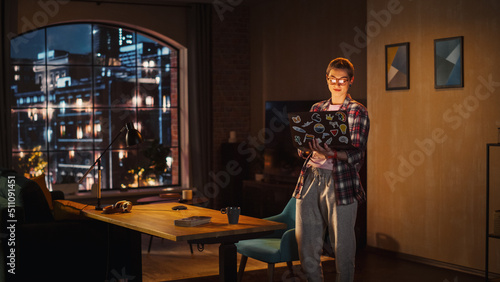 Young Beuatiful Women Working from Home, Standing And Holding Laptop Computer in Stylish Loft Apartment in the Evening. Creative Female Wearing Cozy Casual Outfit. Urban City View from Big Window.