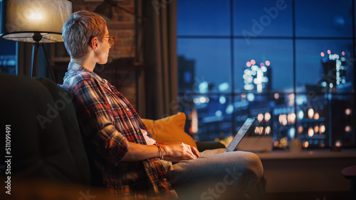 Young Beautiful Woman Sitting on Couch, Using Laptop Computer in Stylish Loft Apartment in the Evening. Creative Female Smiling, Checking Social Media, Stretching. Urban City View from Big Window.