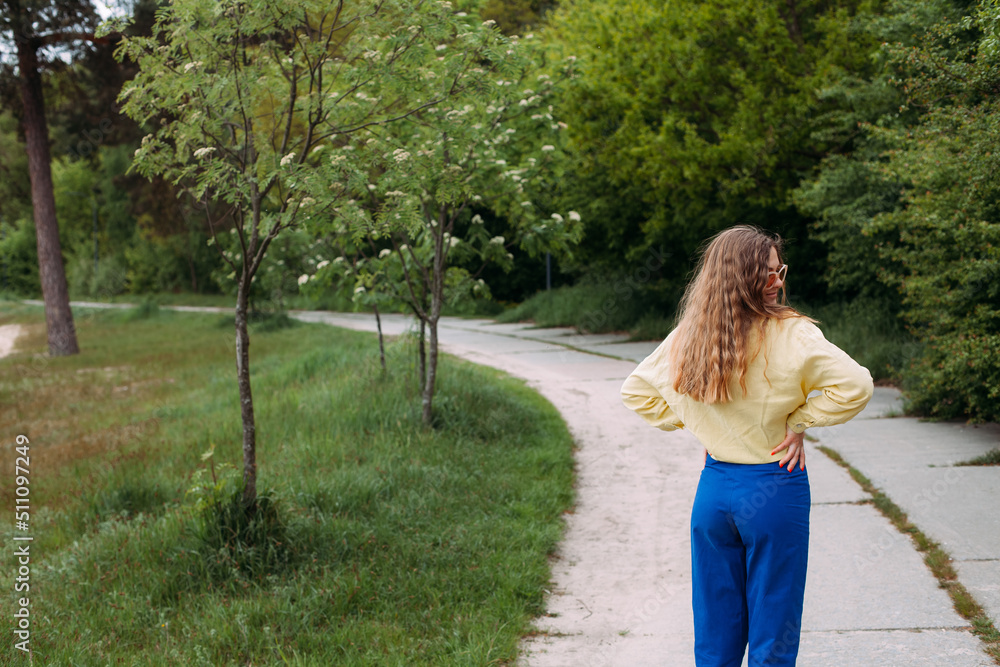 Naklejka premium A smilling girl with blond curly hair dressed in blue jeans and a yellow shirt, wearing sunglasses, stands on a path in a picturesque park in summer.
