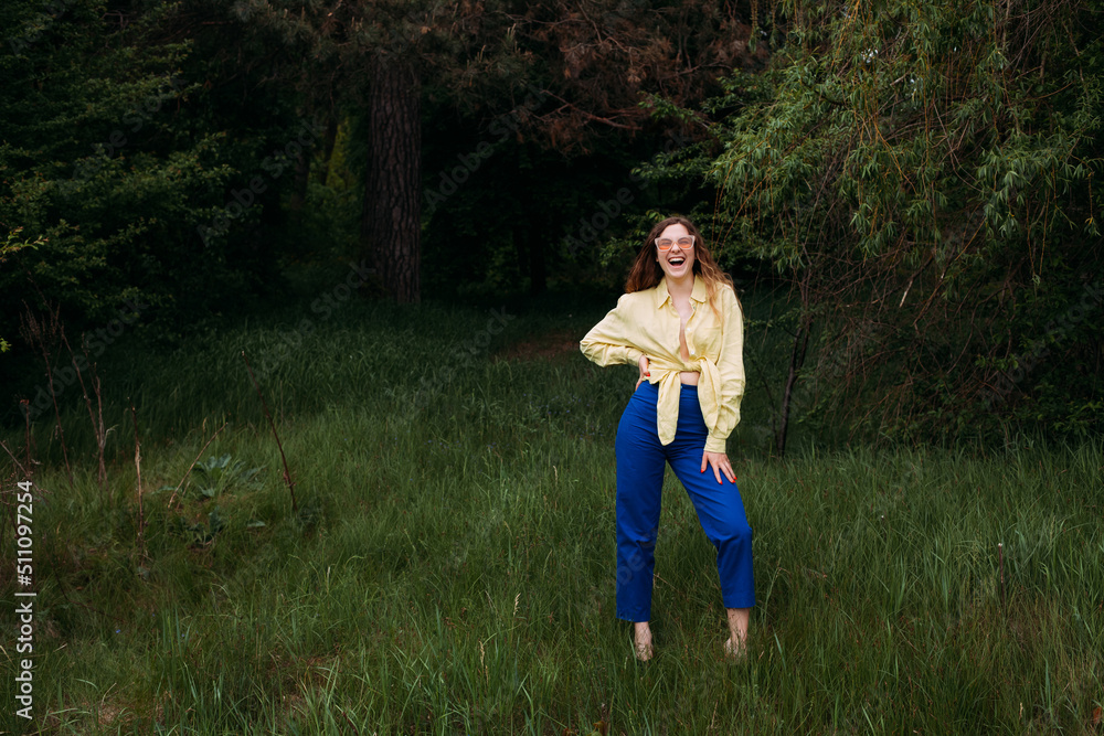 A smilling girl with blond curly hair dressed in blue jeans and a yellow shirt, wearing sunglasses, stands in a picturesque park in summer. A smilling girl with blond curly hair dressed in blue jeans and a yellow shirt, wearing sunglasses, stands in a picturesque park in summer.
