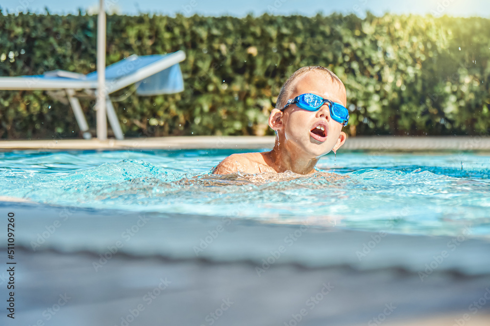 Happy little boy in goggles swimming in blue clean water of outdoor swimming pool during summer holidays in resort