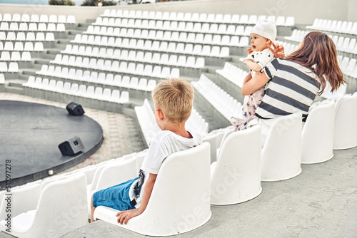 Young mother with sons seats in empty amphitheate rwith white plastic seats for evening show at hotel. Schoolboy shows performance for family photo