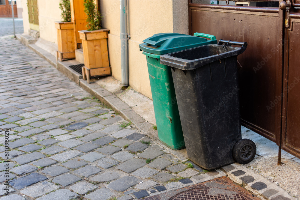 Green and black trash bins next to entrance of a residential home on a paved road with cobblestone Green and black trash bins next to entrance of a residential home on a paved road with cobblestone