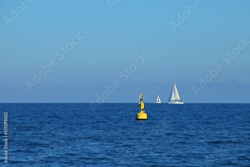 Barcos de vela navegando en el horizonte en la playa de la Malvarrosa en Valencia, España. En primer plano una boya amarilla flotando en medio del mar Mediterráneo de un azul intenso.