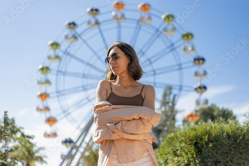 Cheerful smiling girl in sunglasses in front of the Ferris wheel. It's a bright, sunny summer day. 