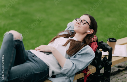 Young beautiful natural looking student lying on a banch in a campus listening to music.