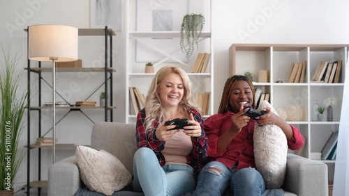 Fun entertainment. Friends home leisure. Weekend joy. Excited girl cheering up bored bff offering to play video game together on couch in modern living room interior. photo
