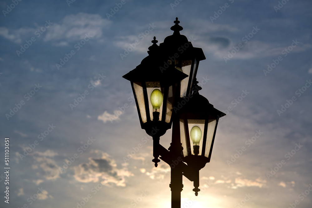 silhouette retro style triple lantern head with transparent glass panels with glow electric light bulbs, iron street lighting in the evening at sunset against a blue sky with sun light.