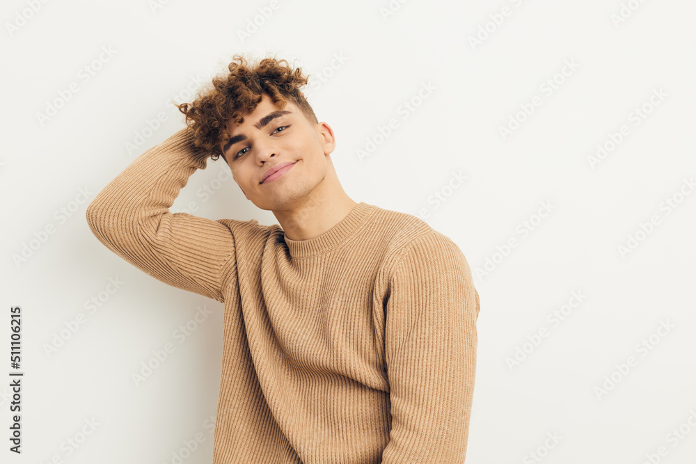 a cute, stylish, curly-haired man stands on a light background looking pleasantly at the camera, in a beige sweater, holding his hands behind his head a cute, stylish, curly-haired man stands on a light background looking pleasantly at the camera, in a beige sweater, holding his hands behind his head