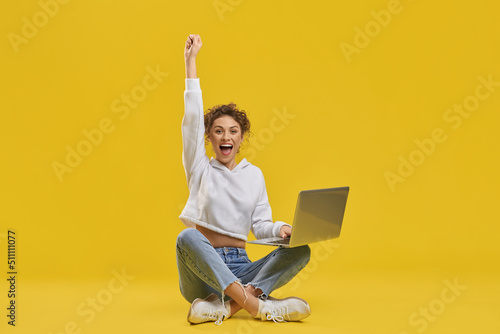Excited woman celebrating victory, looking at netbook screen inside. Front view of happy female freelancer enjoying successful startup, raising hand, isolated on orange background. Concept of emotion. © serhiibobyk