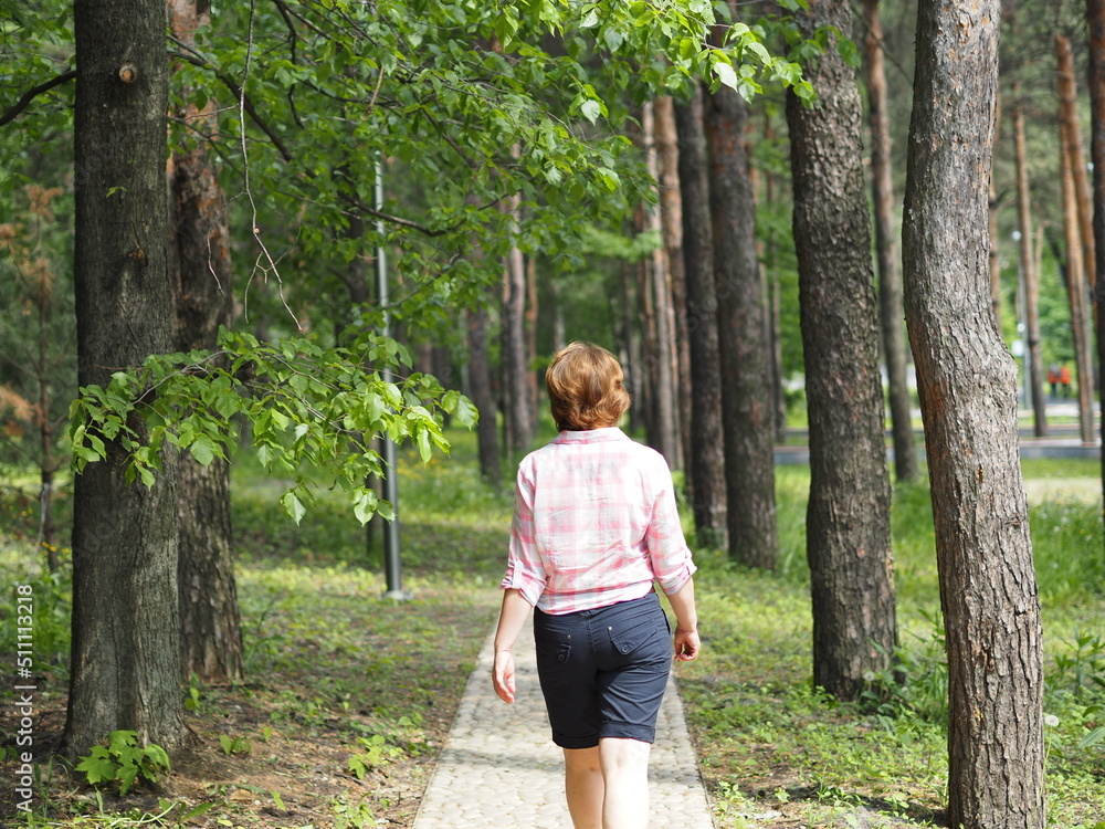 A woman walks in the summer in the park in sunny weather, she walks along a forest path. Back view. A woman walks in the summer in the park in sunny weather, she walks along a forest path. Back view.