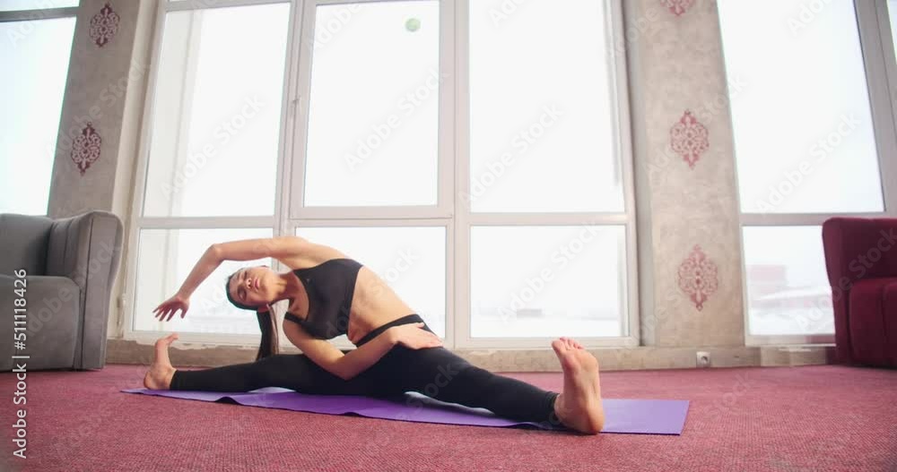 Front view of pretty, brunette lady with ponytail sitting on twine on yoga mat in apartment. Flexble girl bending, stretching, doing yoga alone. Concept of new age reality. Front view of pretty, brunette lady with ponytail sitting on twine on yoga mat in apartment. Flexble girl bending, stretching, doing yoga alone. Concept of new age reality.
