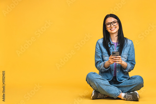 Business concept. Portrait of happy young woman in casual sitting on floor in lotus pose and holding mobile phone isolated over yellow background.