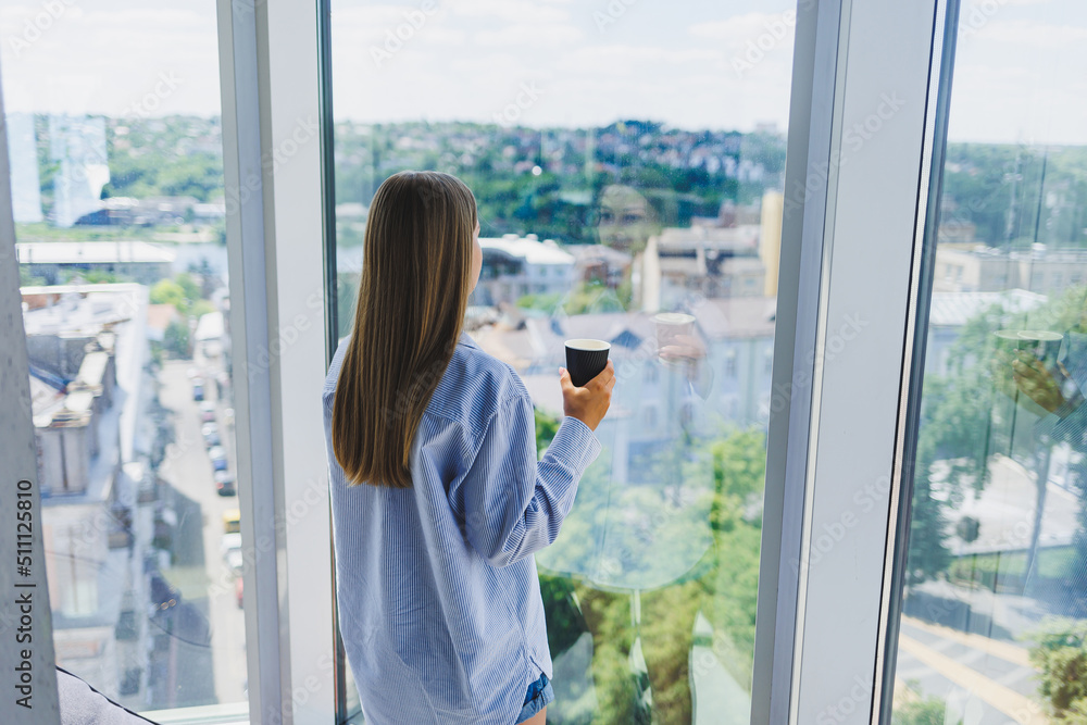 Happy woman drinks coffee and looks out the window in a cafe. A young smiling girl in glasses is standing by the window. Recreation, leisure and free time. The modern freelancer lifestyle