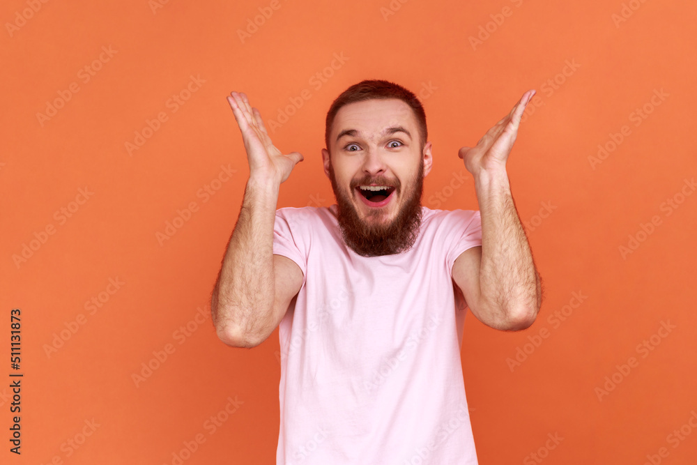 Portrait of bearded man standing with mouth open in surprise, has shocked expression, hears unbelievable news, raised hands, wearing pink T-shirt. Indoor studio shot isolated on orange background. Portrait of bearded man standing with mouth open in surprise, has shocked expression, hears unbelievable news, raised hands, wearing pink T-shirt. Indoor studio shot isolated on orange background.