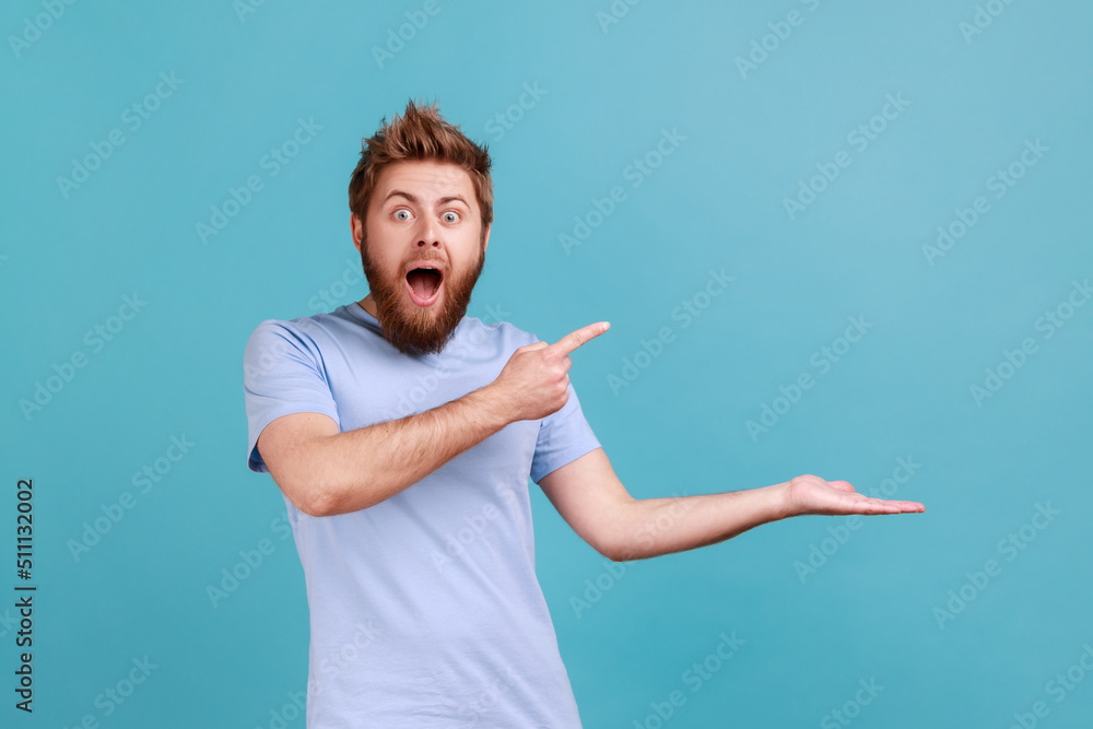 Wow, product advertisement. Portrait of amazed bearded man presenting copy space on palm, showing empty place for commercial text or goods. Indoor studio shot isolated on blue background. Wow, product advertisement. Portrait of amazed bearded man presenting copy space on palm, showing empty place for commercial text or goods. Indoor studio shot isolated on blue background.