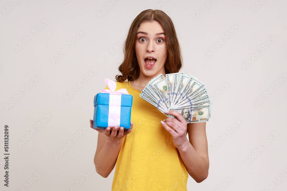 Wow, holiday present shopping! Portrait of amazed teen girl in yellow T-shirt showing dollar banknotes, cashback money and gift box, looking shocked. Indoor studio shot isolated on gray background. Wow, holiday present shopping! Portrait of amazed teen girl in yellow T-shirt showing dollar banknotes, cashback money and gift box, looking shocked. Indoor studio shot isolated on gray background.