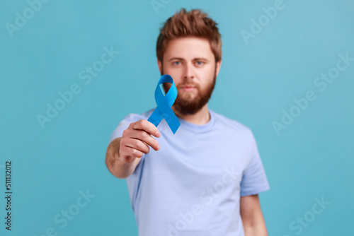 Portrait of serious young adult attractive bearded man holding out blue awareness, disease symbol, looking at camera, support and care. Indoor studio shot isolated on blue background.