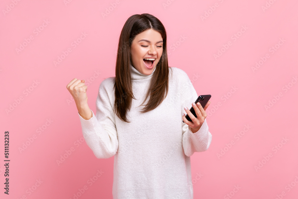 Happy positive girl holds smart phone in hands, clenching fist, celebrating victory, sudden success, wearing white casual style sweater. Indoor studio shot isolated on pink background. Happy positive girl holds smart phone in hands, clenching fist, celebrating victory, sudden success, wearing white casual style sweater. Indoor studio shot isolated on pink background.