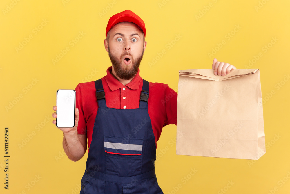 Amazed surprised deliveryman holding paper parcel and showing mobile phone with empty display for advertisement, looking at camera with open mouth. Indoor studio shot isolated on yellow background.