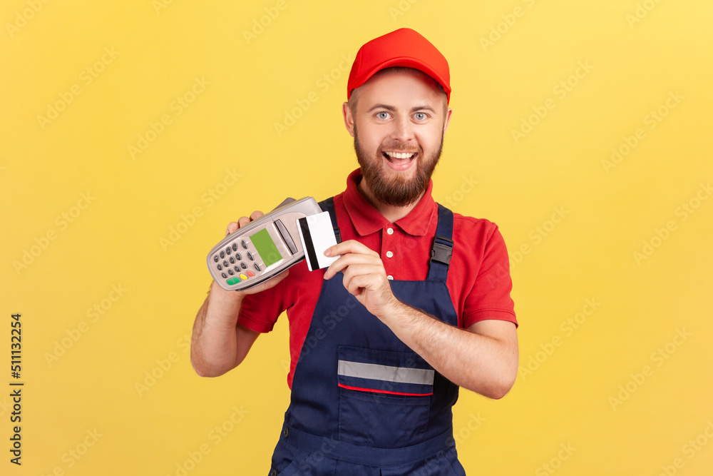 Excited joyful bearded worker man holding pos terminal for contactless payment and credit card, paying for service, expressing happiness. Indoor studio shot isolated on yellow background. Excited joyful bearded worker man holding pos terminal for contactless payment and credit card, paying for service, expressing happiness. Indoor studio shot isolated on yellow background.