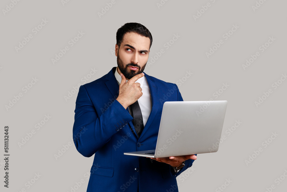 Thoughtful bearded man holding laptop and thinking over startup strategy with serious doubtful expression, wearing official style suit. Indoor studio shot isolated on gray background. Thoughtful bearded man holding laptop and thinking over startup strategy with serious doubtful expression, wearing official style suit. Indoor studio shot isolated on gray background.