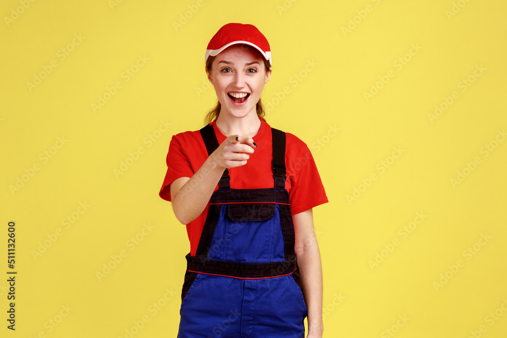 Portrait of excited happy worker woman standing and pointing at camera, laughing loud out, hearing funny joke, wearing overalls and red cap. Indoor studio shot isolated on yellow background. Portrait of excited happy worker woman standing and pointing at camera, laughing loud out, hearing funny joke, wearing overalls and red cap. Indoor studio shot isolated on yellow background.
