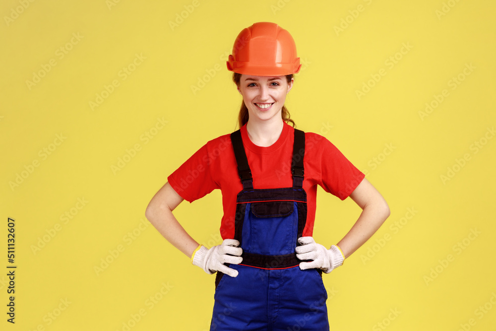 Portrait of smiling confident builder woman standing holding hands on hips, looking at camera, wearing overalls, gloves and protective helmet. Indoor studio shot isolated on yellow background. Portrait of smiling confident builder woman standing holding hands on hips, looking at camera, wearing overalls, gloves and protective helmet. Indoor studio shot isolated on yellow background.