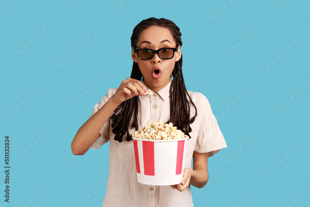 Excited woman with dreadlocks in 3d imax glasses watching movie film, hold popcorn, watching interesting movie, keeping mouth open, wearing white shirt. Indoor studio shot isolated on blue background. Excited woman with dreadlocks in 3d imax glasses watching movie film, hold popcorn, watching interesting movie, keeping mouth open, wearing white shirt. Indoor studio shot isolated on blue background.