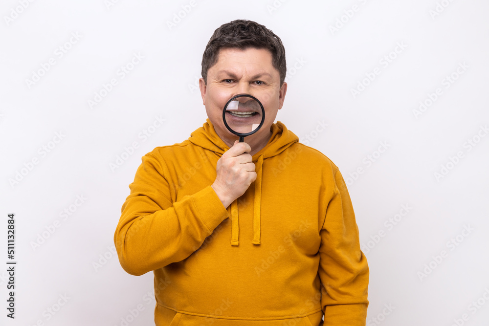 Portrait of middle aged man standing, holding magnifying glass on his teeth, looking with toothy smile, wearing urban style hoodie. Indoor studio shot isolated on white background. Portrait of middle aged man standing, holding magnifying glass on his teeth, looking with toothy smile, wearing urban style hoodie. Indoor studio shot isolated on white background.