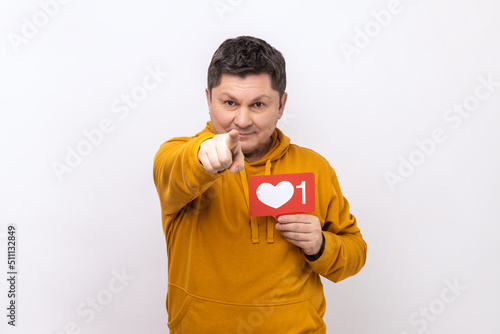 Portrait of happy positive man showing blogger heart icon and pointing to camera, asking to click button, wearing urban style hoodie. Indoor studio shot isolated on white background.