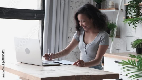 African American woman sits indoor and using app on laptop for video call to friends, making notes in a notebook. Young female employee in casual wear talking online via video connection waving hands photo