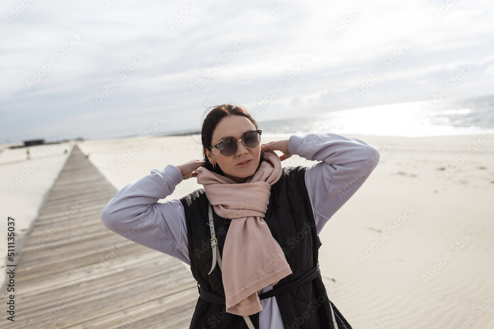 Fashionable pretty woman with vintage eyewear in casual clothes with scarf, vest and sweatshirt walks on wooden walkways on the sandy beach near a ocean