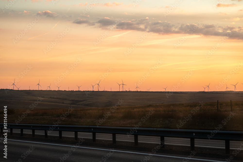 Many windmills stand in endless field and generate environmental energy on horizon in orange and yellow color. Long road goes into distance against backdrop of windmills against backdrop of setting.